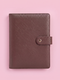 A brown textured leather planner cover with a snap-button strap closure, laid flat on a solid pink background.