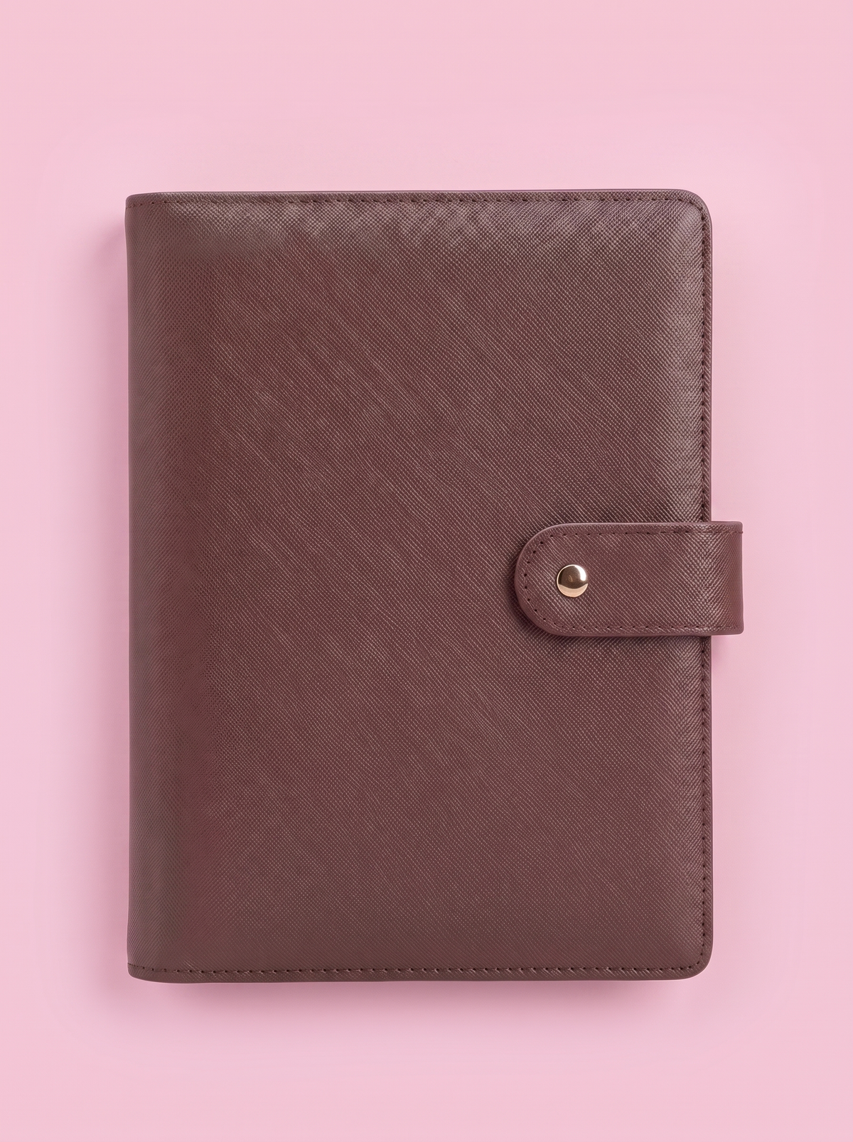 A brown textured leather planner cover with a snap-button strap closure, laid flat on a solid pink background.